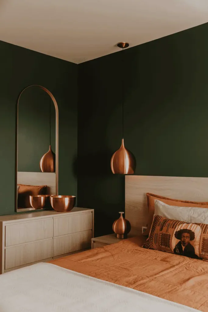 Modern bedroom with deep forest green or charcoal walls, light ash or blonde oak furniture, copper accents throughout including copper bowls, mirror frame, and pendant lights. African print pillows in warm tones bridging dark walls and light wood. Contemporary Afrohemian aesthetic, warm copper glow, editorial interior photography with unexpected modern color palette.