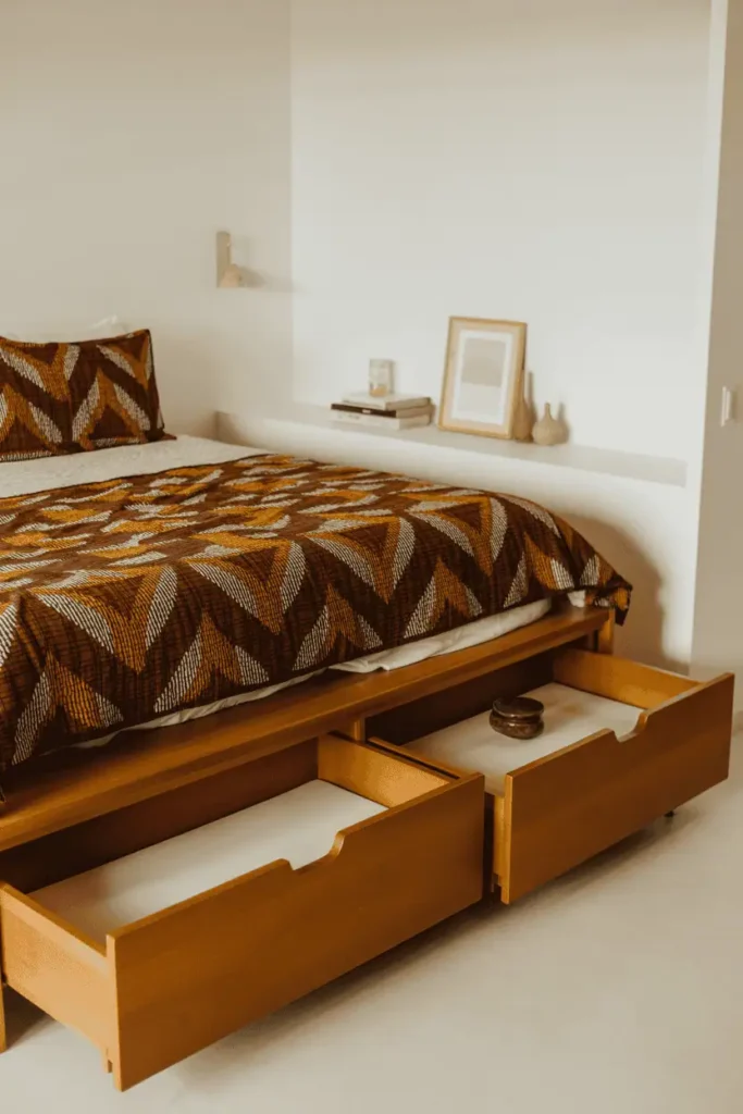 Functional platform bed with built-in drawers underneath in medium-toned solid wood, bold geometric African wax print bedding on top. White walls, floating shelf with minimal objects, clean and uncluttered space. Practical yet stylish aesthetic, bright natural lighting, editorial interior photography showing smart storage solutions with cultural textile elements.