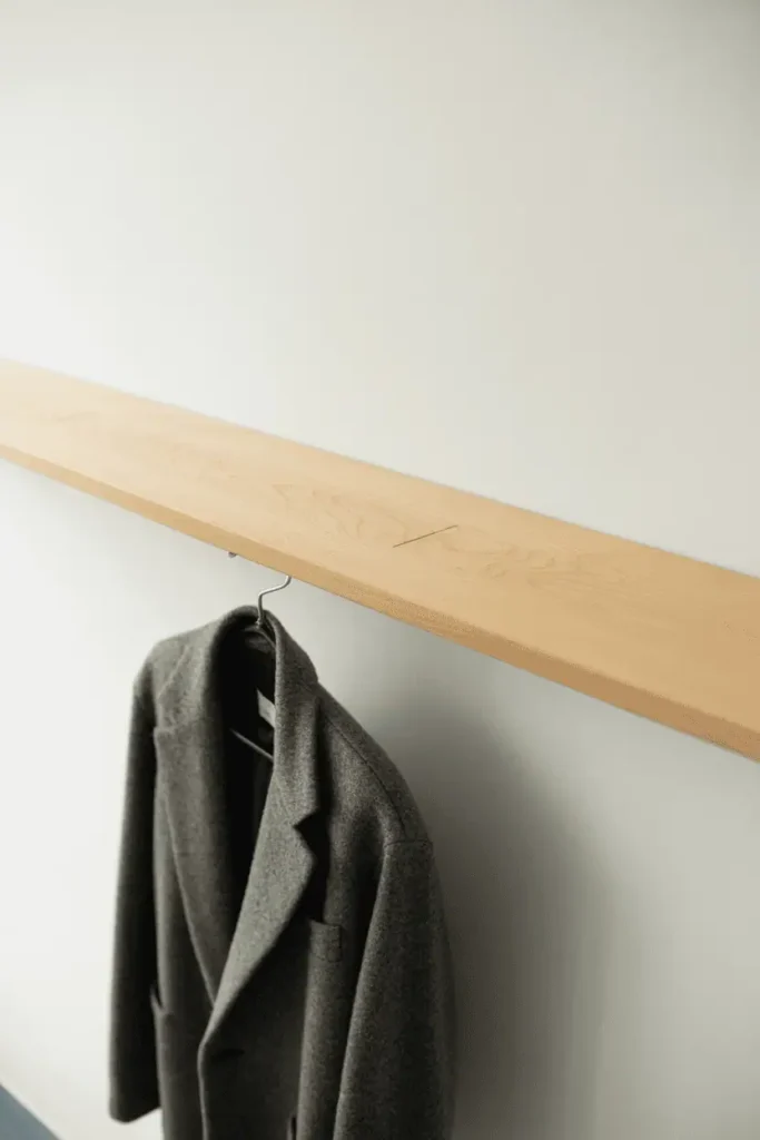 A sleek horizontal maple wood bar appearing to float off a clean white wall with no visible mounting hardware. One charcoal gray wool coat hangs centered on the bar. Bright, airy lighting with minimal shadows. High-key aesthetic, extremely clean and modern. Architecture magazine style, precise and minimal.