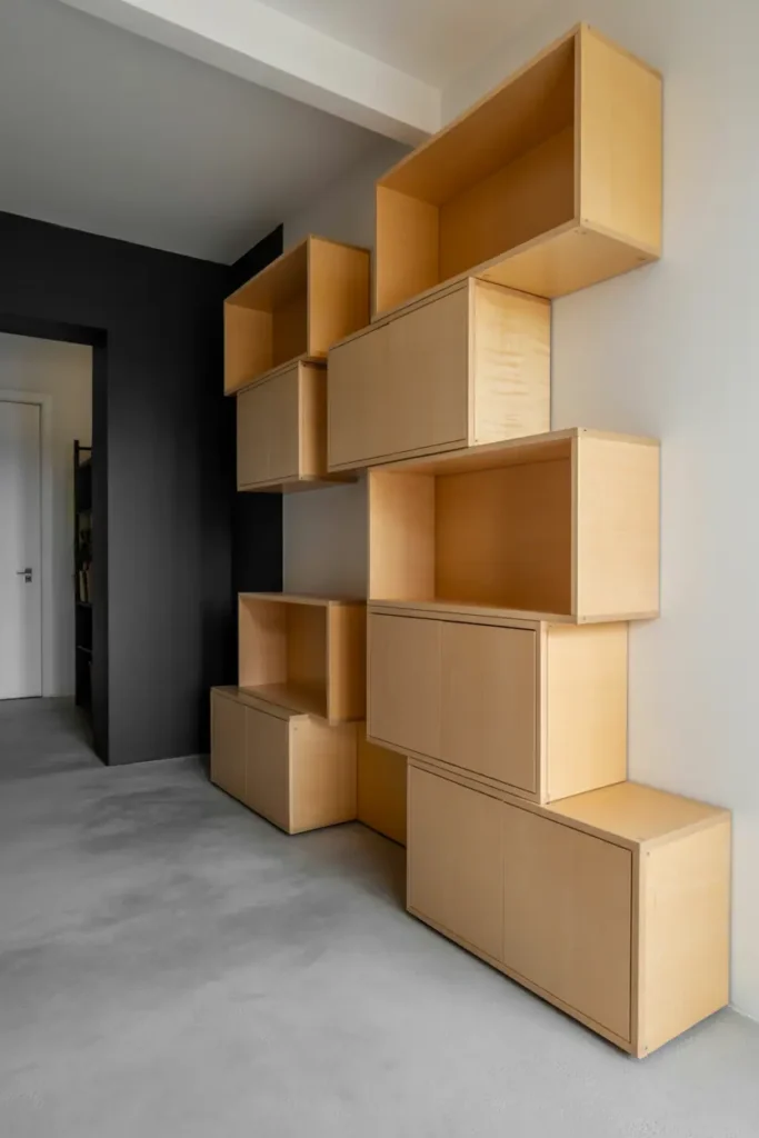 Contemporary hallway with asymmetric modular birch shelving units arranged on wall. Different sized boxes in balanced configuration - some open, some with doors. Natural birch finish. Polished concrete floor, charcoal painted opposite wall. Modern functional design. Clean lighting, contemporary aesthetic, interior photography.