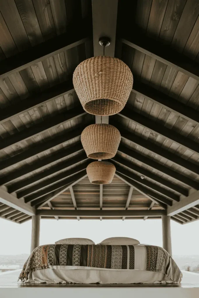 Bedroom with exposed dark-stained wooden ceiling beams, woven seagrass pendant lights hanging at varying heights. Low simple bed below, mudcloth and African print textiles layered on bed. High ceiling lofted space, architectural focus on ceiling elements, natural light from windows, editorial interior photography emphasizing vertical space and architectural features.