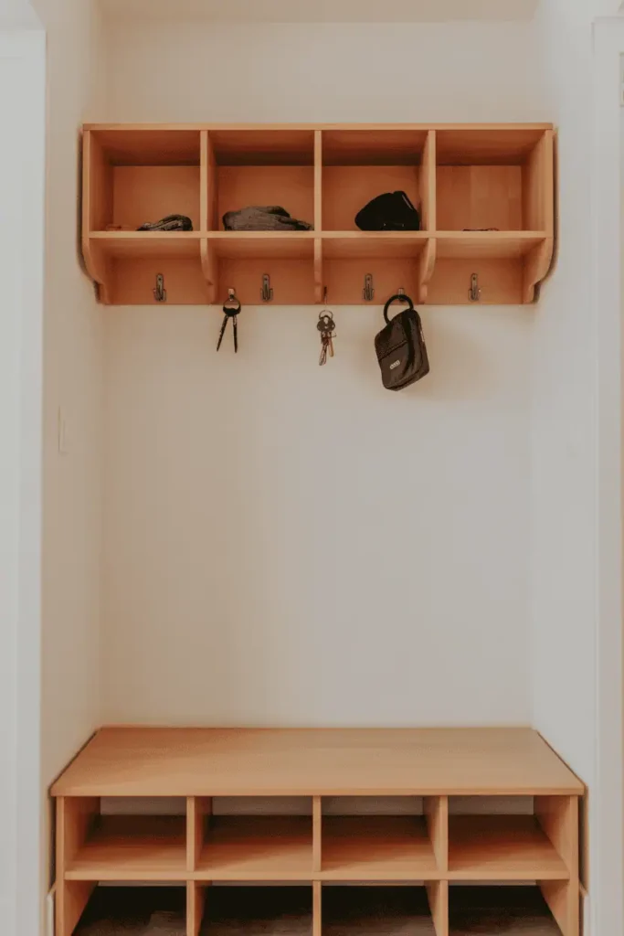 Small entryway with narrow light wood bench and wall-mounted shelf above divided into 4 cubbies for organized storage. Simple bench seat, no under-bench storage, cubbies hold keys and small items. White walls, vertical organization, family-friendly design. Natural lighting, practical and clean aesthetic. Interior photography showing vertical storage efficiency, approximately 5 feet of wall space.