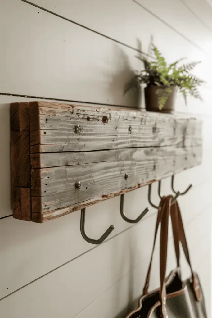 A thick plank of weathered gray reclaimed barn wood mounted as a floating ledge with visible old nail holes and patina. Black metal hooks drop from the underside. A small potted fern sits on top while a leather bag hangs below. White shiplap wall background, soft diffused light. Rustic yet refined, editorial styling with earthy tones.