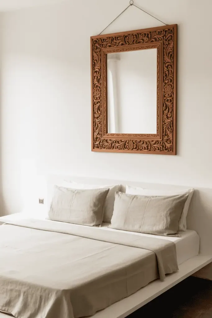 Minimalist Indian bedroom with white walls, pale wood platform bed, natural linen bedding. Single large carved wooden mirror frame in teak with traditional floral and geometric carving hanging above bed. The mirror is the only ornate element. Clean, intentional, contemporary with cultural reference. Soft natural light, editorial photography.