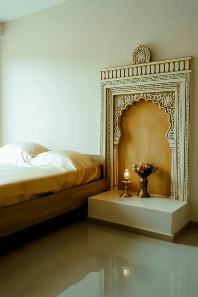 Indian bedroom with small carved wooden mandir prayer alcove built into wall beside bed, painted soft gold and white with carved details. Minimal bedroom: simple wooden bed, white walls, polished floor. Brass lamp in mandir, fresh flowers. Spiritual and personal element integrated architecturally. Warm lighting, intimate, cultural detail.