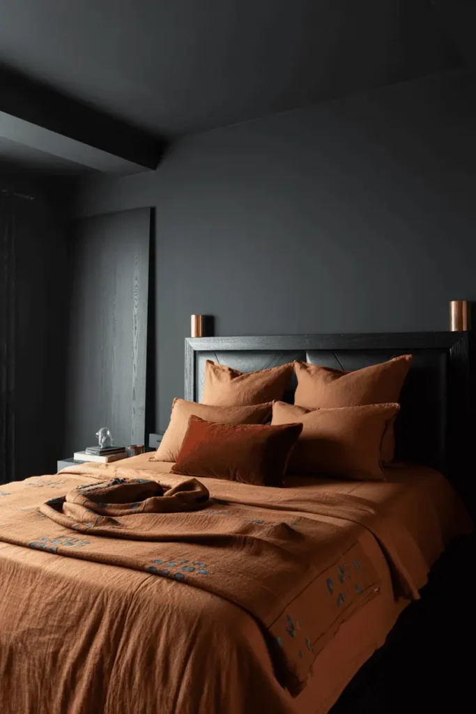 Dramatic bedroom with charcoal gray walls, rust and burnt orange bedding, dark espresso wooden furniture, brass accents catching light against dark walls. Large mudcloth throw at foot of bed. Moody and cocooning atmosphere, combination of ambient and focused lighting, window with bright daylight for contrast, editorial interior photography with dramatic lighting.