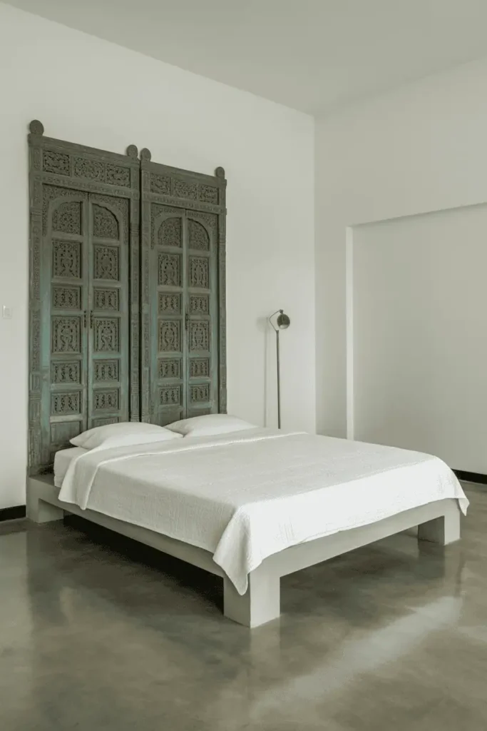 Minimalist bedroom with pair of vintage Indian carved doors in faded teal mounted horizontally as headboard. Tall, intricate carving, positioned above simple low platform bed with white bedding. Concrete floors, white walls, single brass floor lamp. Contemporary setting with traditional artifact. Clean, intentional, editorial style photography.