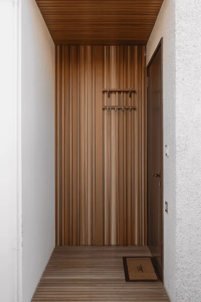 Very narrow hallway entryway with vertical wood slat accent wall in medium tone running floor to ceiling, three simple metal hooks attached to slats. Opposite wall painted white, completely clear floor with only doormat. Architectural wood detail, shadow play from slats, natural lighting. Professional interior photography emphasizing vertical space and texture without furniture.
