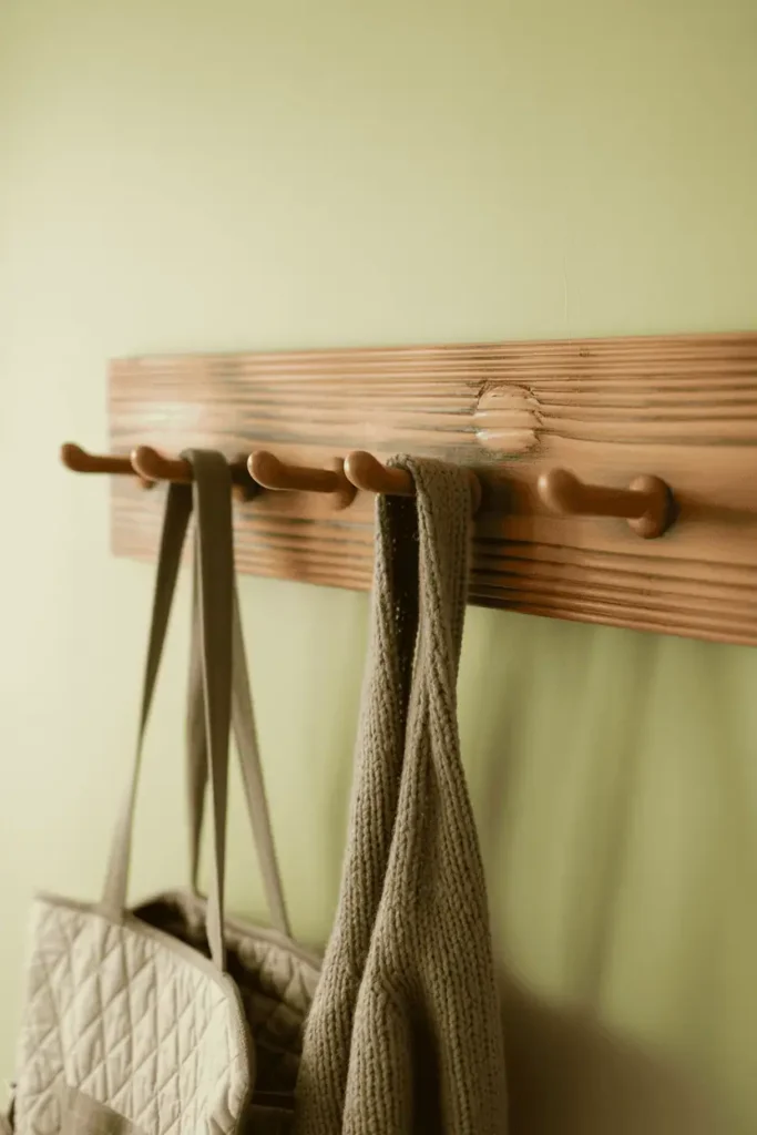 A pine peg rail with warm antique brown stain showing subtle distressing in the grain, mounted on a soft cream cottage-style wall. Four pegs with gentle vintage character. A quilted bag and knit scarf hang casually. Soft diffused natural light, cozy and lived-in feel with vintage warmth.