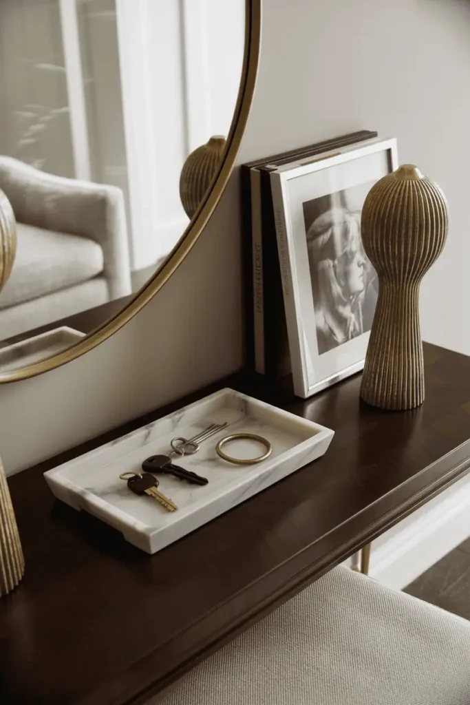 Upscale entryway with dark mahogany console table styled with luxury materials, marble tray holding keys and objects, tall sculptural brass lamp, large art book standing upright. Oversized round brass-framed mirror above. Neutral palette of whites, grays, brass, dark wood. Professional interior photography, elegant and refined aesthetic, high-end materials.