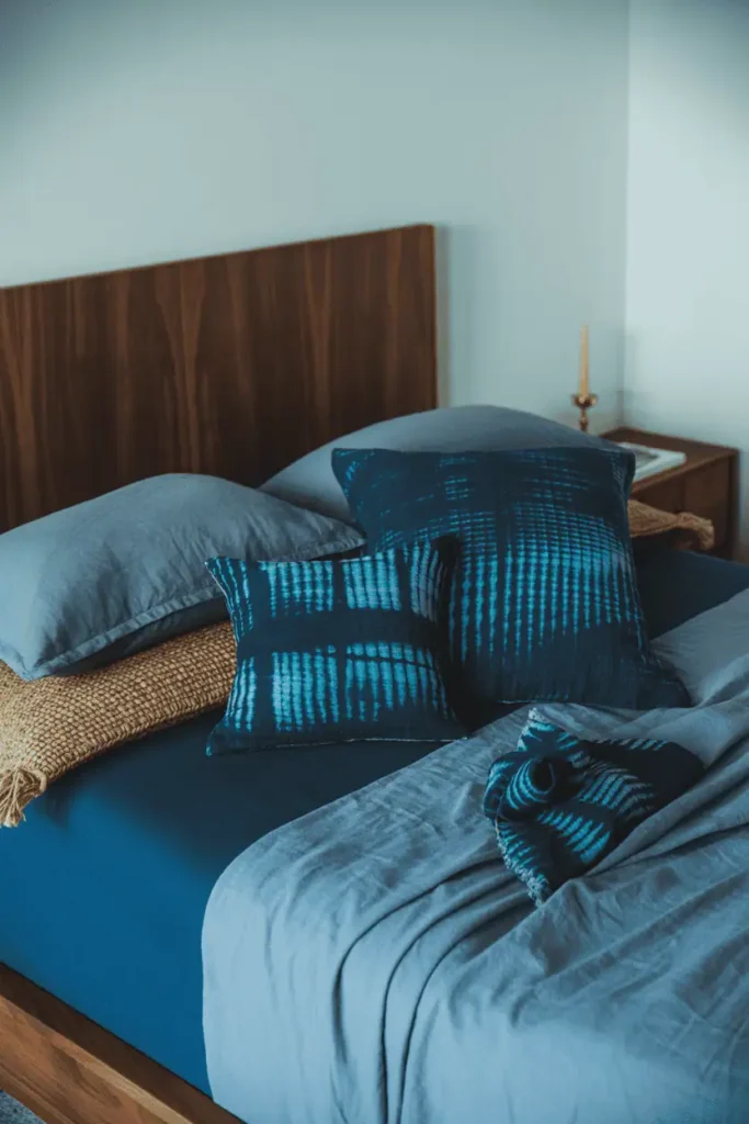 Bedroom with dark walnut furniture, indigo-dyed textiles and navy mudcloth bedding, deep blue wax print accents. White walls, natural jute elements, single brass candleholder. Cool blue tones with warm wood contrast, calm and slightly formal aesthetic, soft natural lighting, editorial interior design photography with cooler color palette.