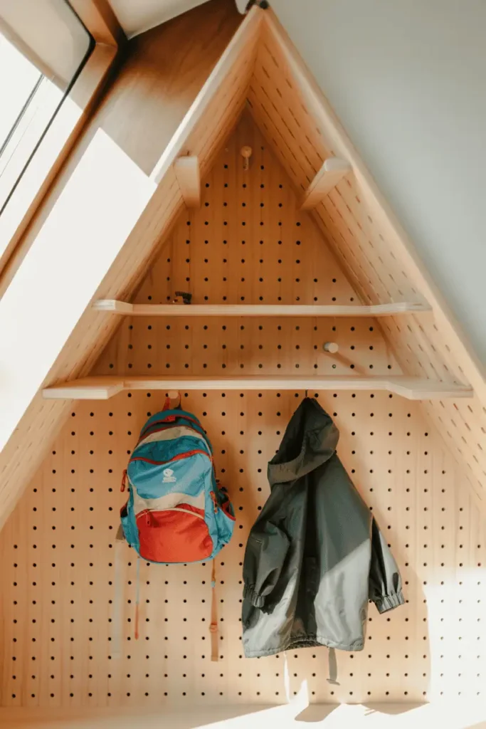 A diagonal view into a room corner showing a triangular wooden peg board in natural ash that fits perfectly into the 90-degree angle. Pegs radiate outward at various points. A child's colorful backpack and rain jacket hang on lower pegs. Natural window light from the side, clever space-saving design with warm family-friendly feel.