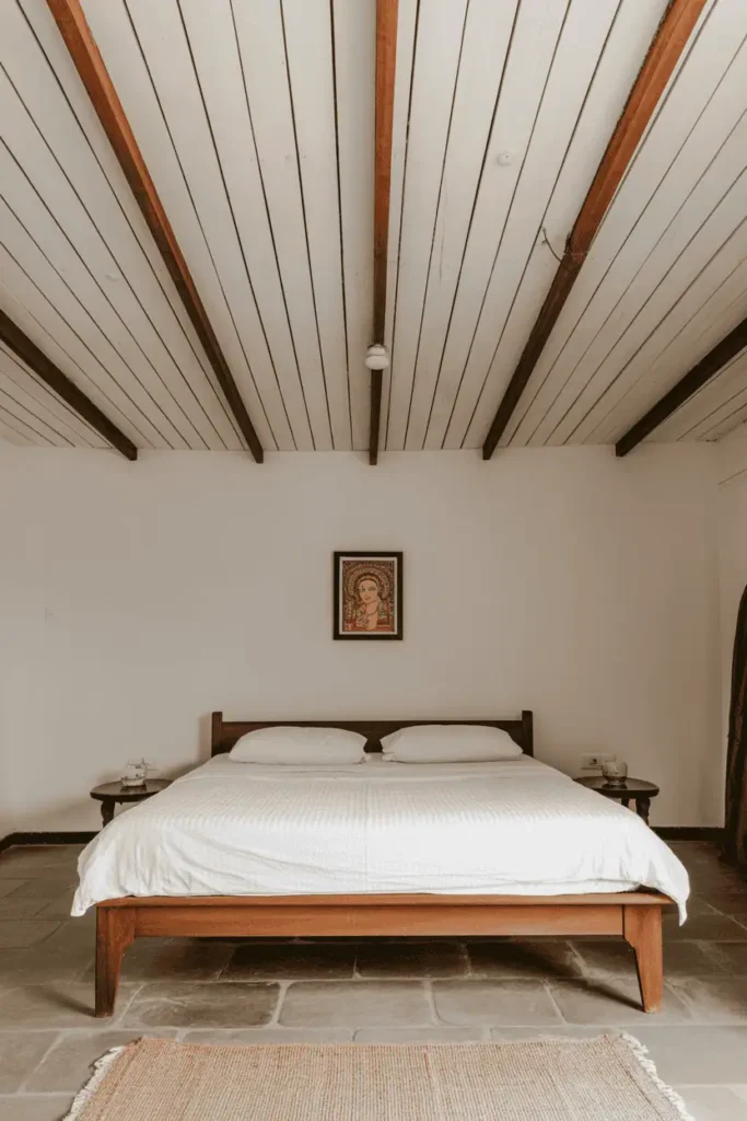 Minimalist Indian bedroom dominated by whitewashed wood plank ceiling with darker exposed beams running perpendicular. Simple low teak bed frame with white linens below. Single framed Madhubani painting on bare wall. Polished stone floor with large cotton dhurrie. Restful, uncluttered, architectural focus. Natural light, serene atmosphere.