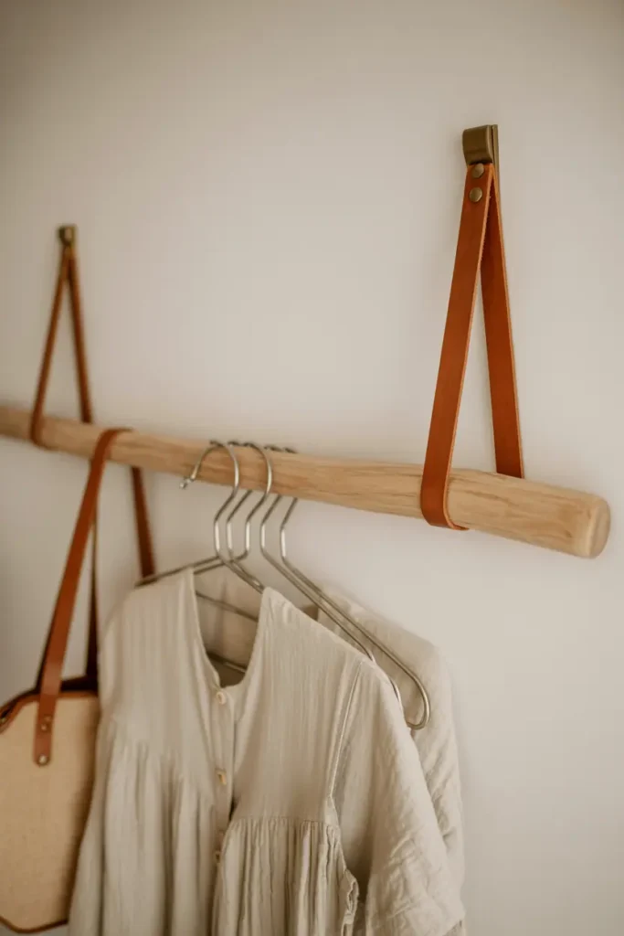 A smooth wooden dowel rod suspended horizontally by tan leather straps from a white wall. The leather straps show natural grain and brass hardware. Several items hang from the rod including a linen dress and tote bag. Soft natural light, handmade aesthetic with warm tactile materials. Bedroom or closet setting.
