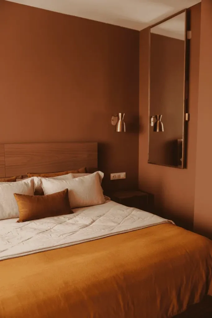 Bedroom with warm terracotta painted walls, medium-toned walnut bed frame, cream and rust bedding, brass wall sconce and mirror frame catching light. Simple wooden nightstand, minimal styling. Warm ambient lighting, rich earth tones, cozy Afrobohemian aesthetic, professional interior photography with attention to color harmony.