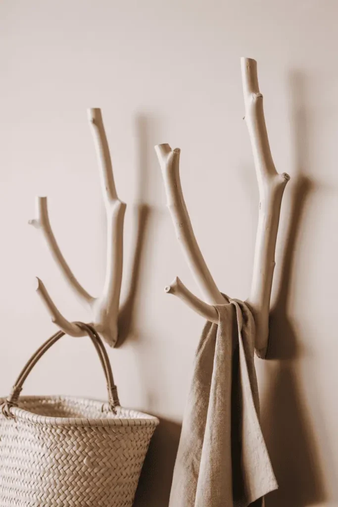 Three smooth sanded branch hooks mounted at different angles on a soft white wall. The branches show natural taper and subtle knots but are refined, not rough. A woven market basket and linen tea towel hang from two branches. Soft diffused natural light, botanical and organic aesthetic with muted earth tones.