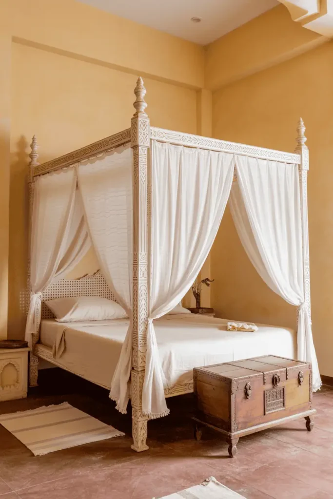 Traditional Indian four-poster bed in pale unstained teak wood, charpai-inspired design. Sheer white cotton fabric draped loosely across posts and sides. Ochre plaster walls, terracotta tile floor with scattered cotton rugs. Carved wooden trunk at foot of bed. Airy, haveli-style bedroom. Warm climate aesthetic, natural light, heritage home feel.