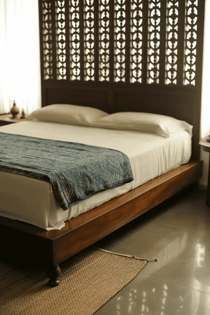 Indian bedroom with low dark sheesham wood platform bed, tall carved jali headboard with geometric cutwork reaching toward ceiling. White cotton bedding, single indigo block-printed quilt. Natural light filtering through jali creating shadow patterns. Polished concrete floor with natural fiber handwoven dhurrie. Minimal brass accessories. Warm natural light, traditional craft, interior design photography.